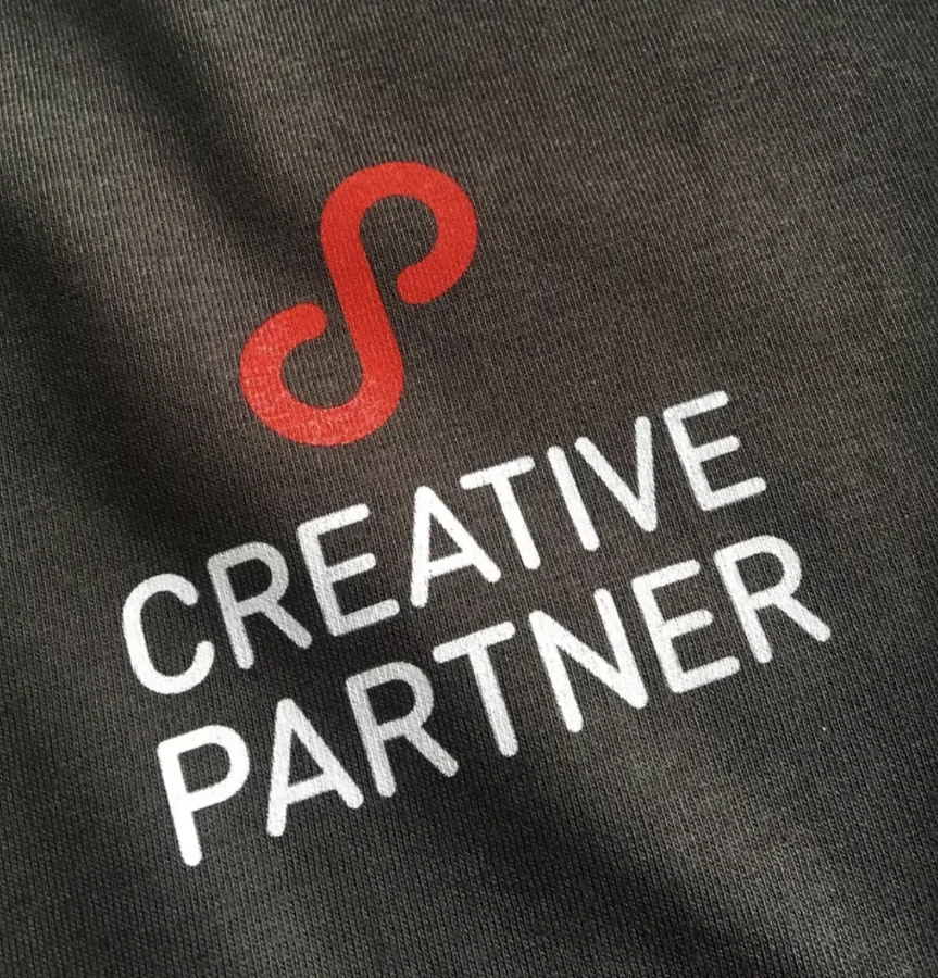 Raudonas ir baltas „Creative Partner“ logotipas ant tamsaus audinio – Teeboxas atliktas darbas